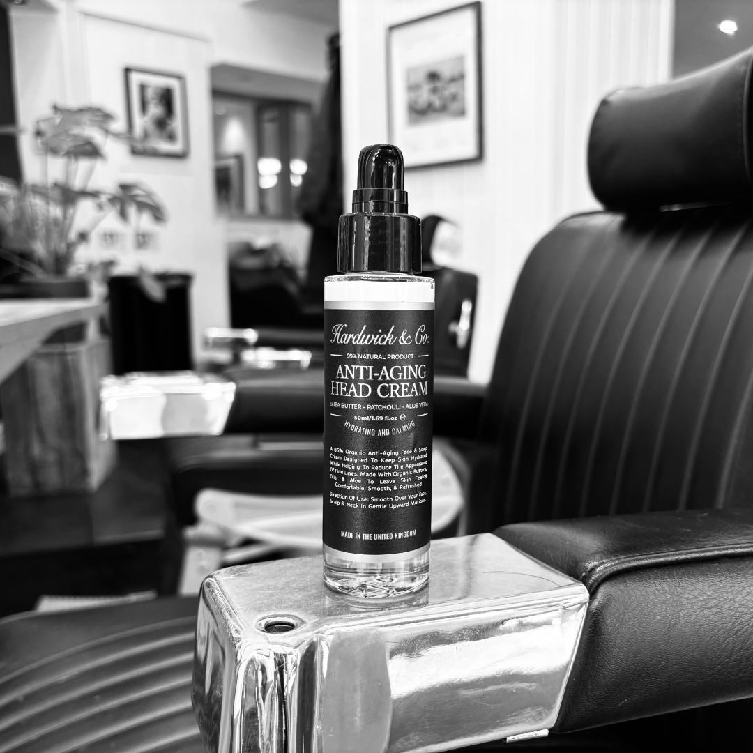 A bottle of Hardwick & Co. Anti-Aging Head Cream stands on a polished metallic surface in a salon setting, with a black leather chair in the background.