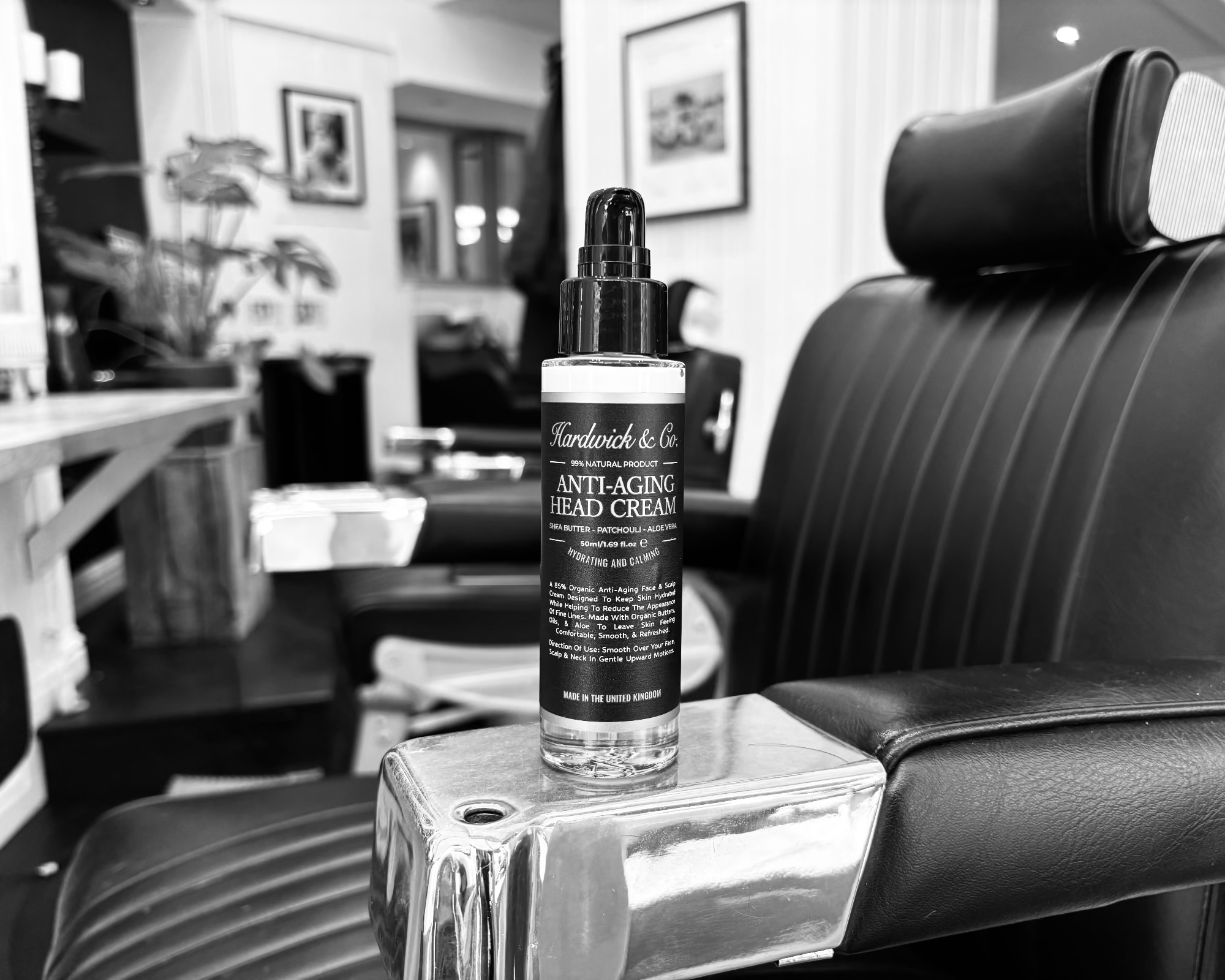A bottle of Hardwick & Co Anti aging Head Cream sits on the chrome armrest of a black leather barber chair in a barbershop.