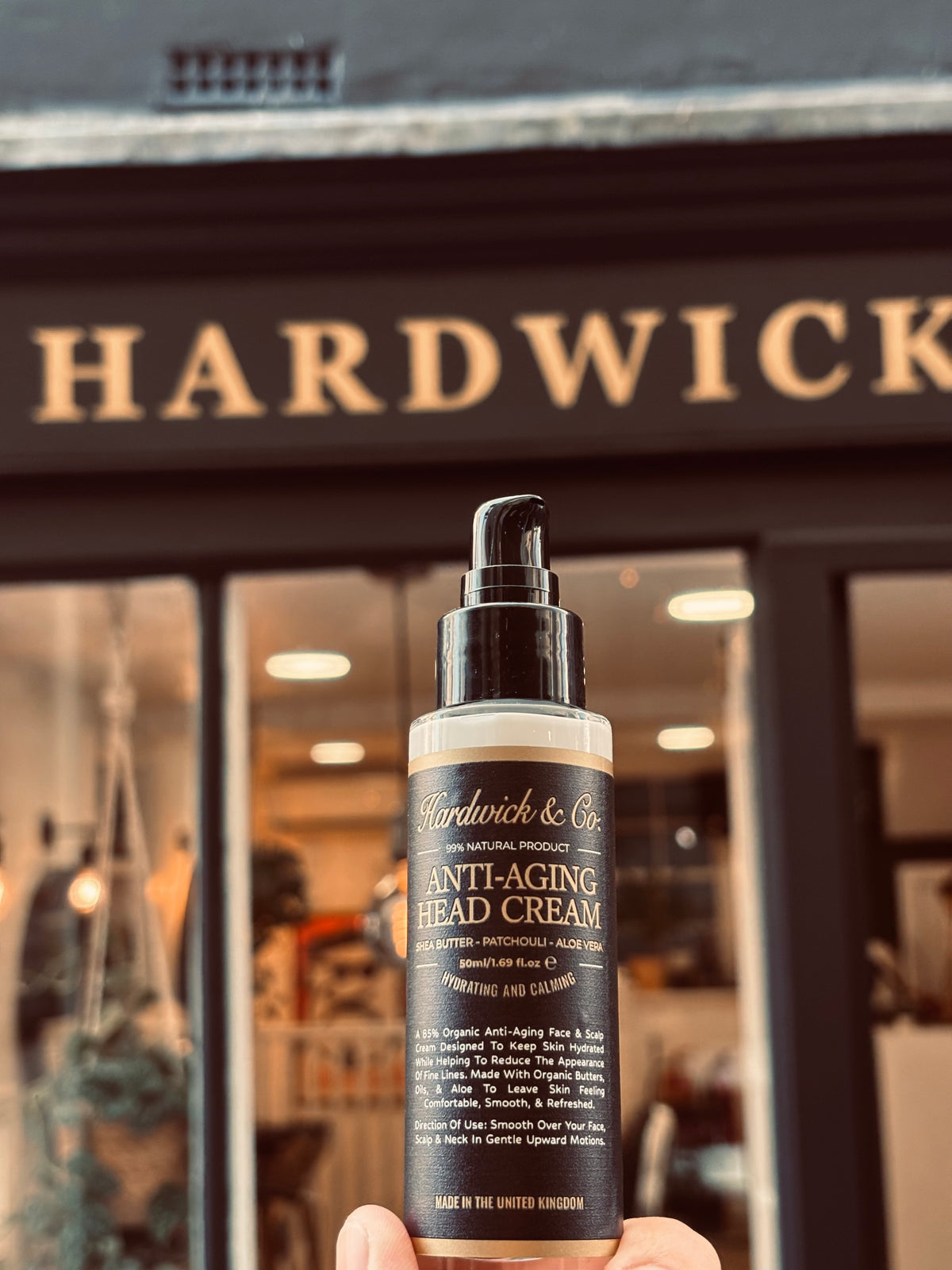 A hand holds a bottle of Hardwick & Co. Anti-Aging Head Cream in front of a blurred HARDWICK storefront.