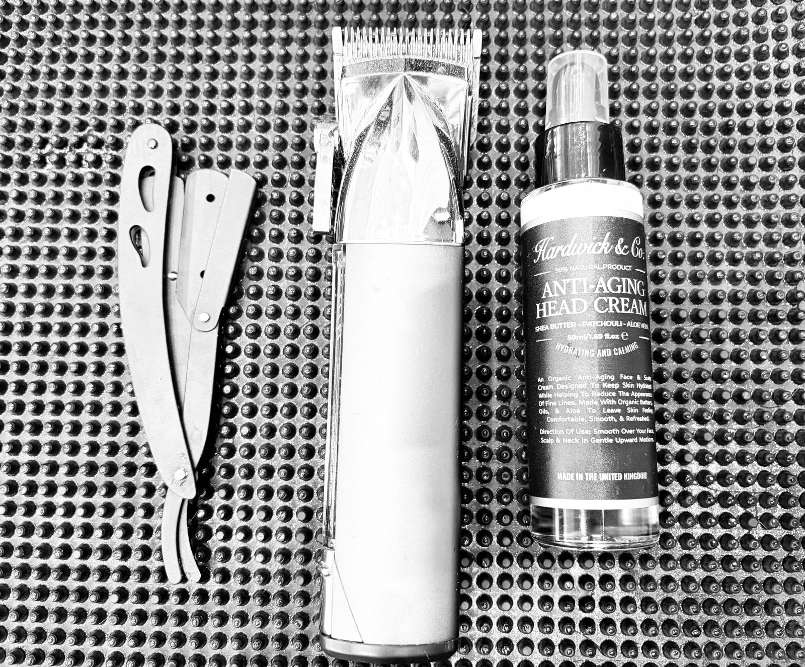 Set of grooming products including a straight razor, hair clipper, and head cream on a textured surface.